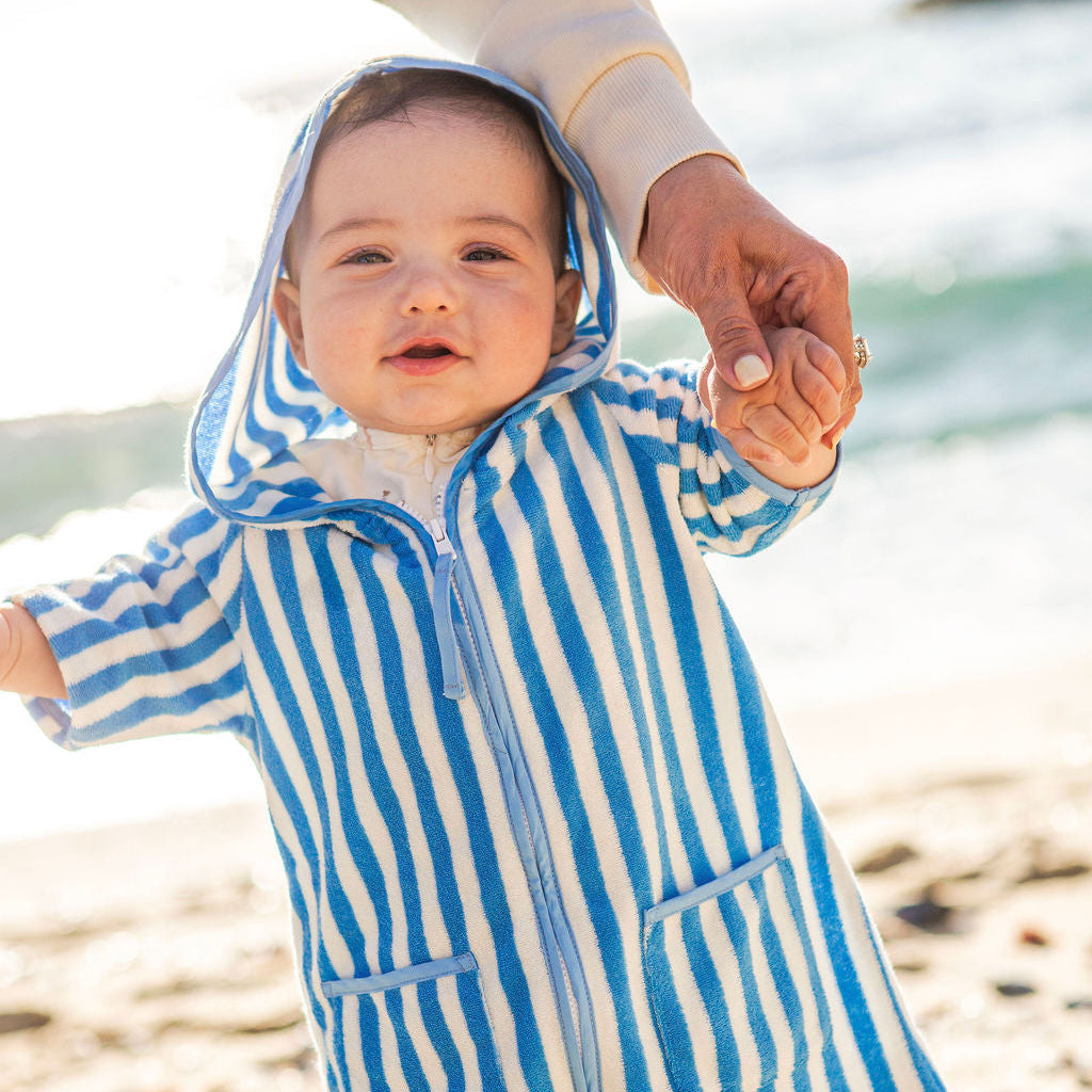 Baby in a blue and white striped beach poncho standing on a beach.