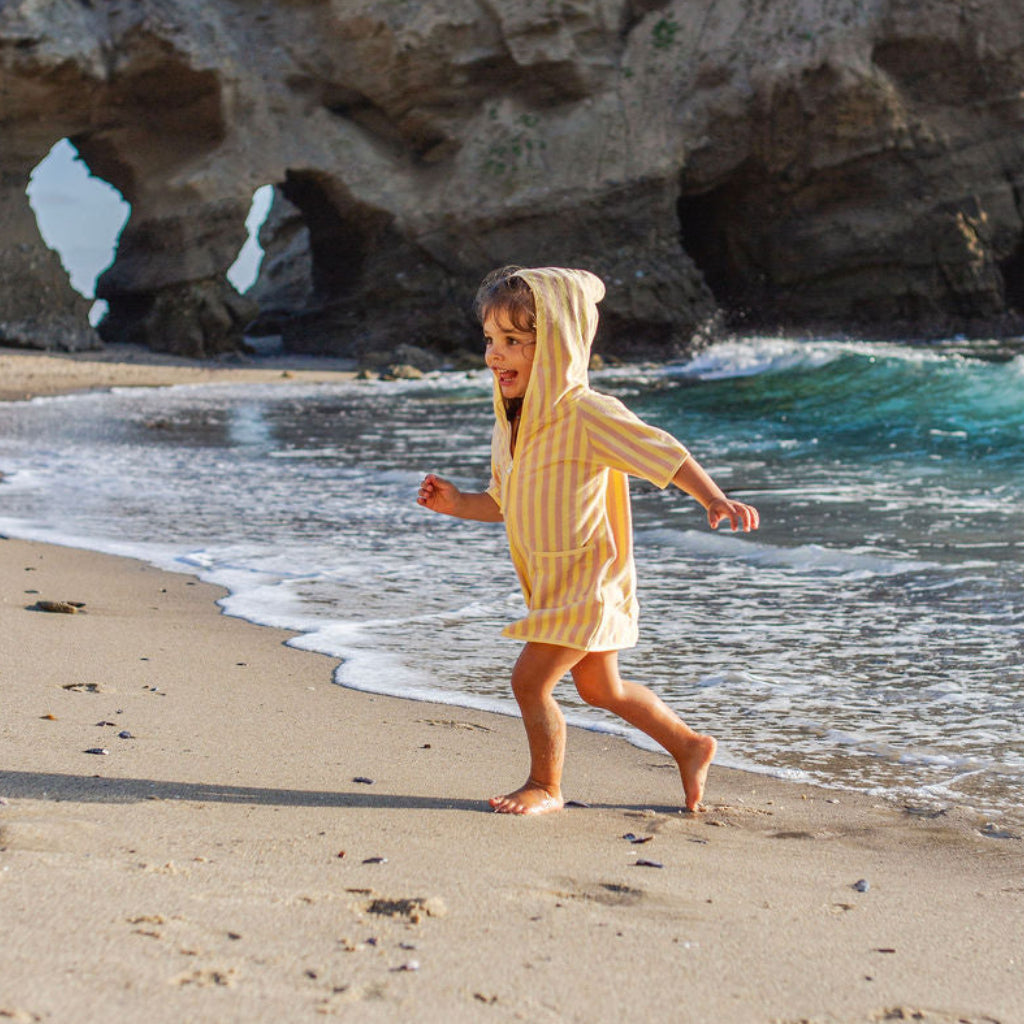 Child in a yellow and pink striped poncho running on a sandy beach with rocky cliffs and ocean waves in the background.