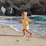 Child in a yellow and pink striped poncho running on a sandy beach with rocky cliffs and ocean waves in the background.