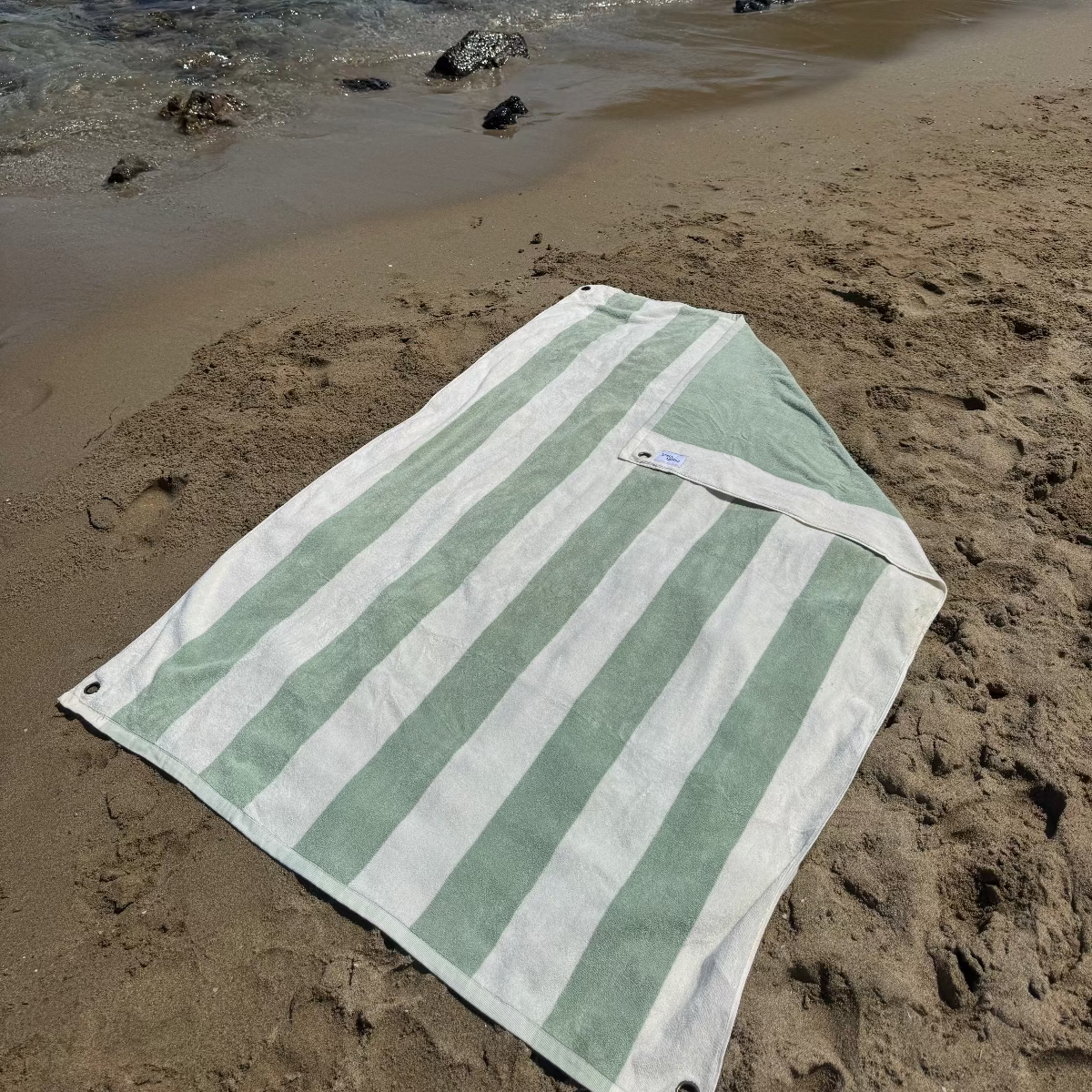 Green and white striped towel on a sandy beach with people and umbrellas in the background.