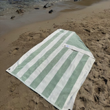 Green and white striped towel on a sandy beach with people and umbrellas in the background.